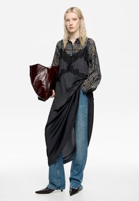 Vestido de satén negro con acentos de encaje superpuesto sobre una camisa de cuadros, combinado con jeans azul claro y un bolso burdeos brillante. Zapatos negros de punta.