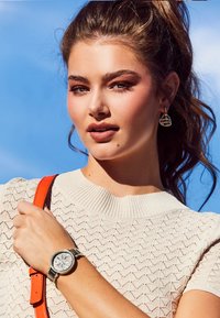 Een lichtbeige, gestructureerde top met een ribbelhalslijn, gecombineerd met een metallic horloge met een gedetailleerde wijzerplaat en een oranje schouderband.