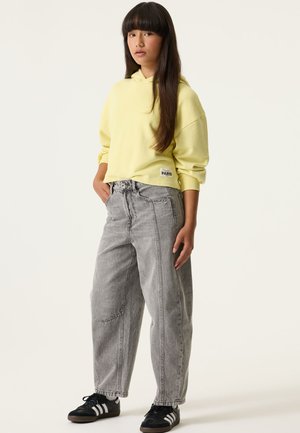 Jonge persoon staat met handen in de zakken, draagt een gele hoodie, losse grijze jeans en zwarte sneakers met witte strepen op een effen achtergrond.