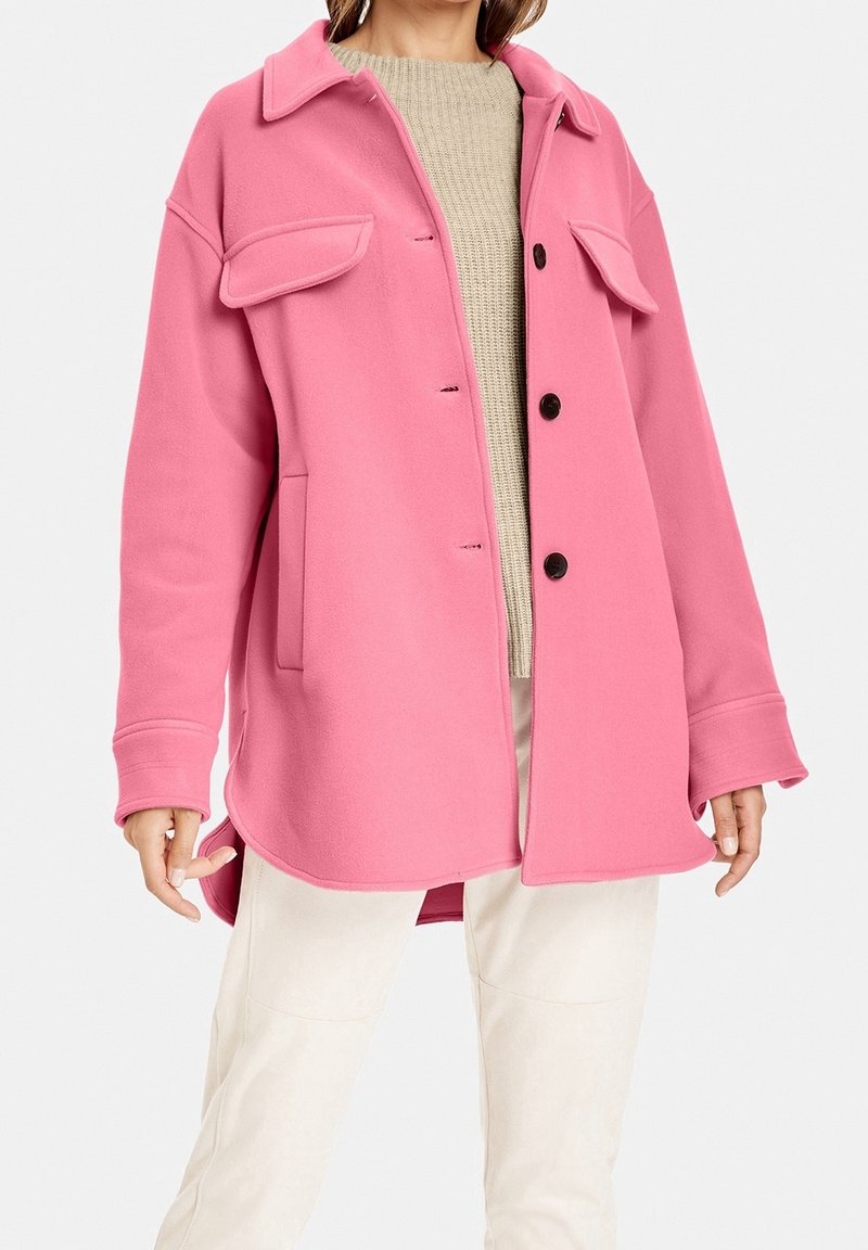 Pink fleecejakke med krave, to forlommer og sorte knapper. Bæres over en beige striktop og cremefarvede bukser.