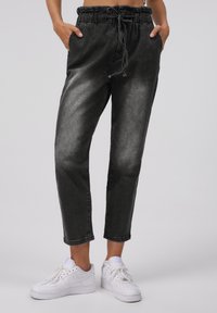 Personne portant un pantalon de jogger noir délavé avec taille élastique et des baskets blanches, les mains dans les poches, debout sur un fond uni.