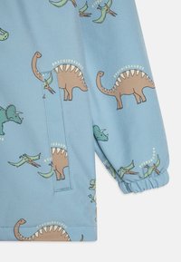 Jasnoniebieska tkanina z wzorem dinozaurów, w tym Brachiozaur i Pteranodon, z elastycznymi mankietami i detalem bocznej kieszeni.
