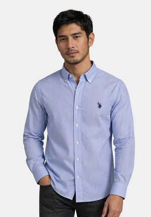 Mann trägt hellblaues gestreiftes Hemd mit Knöpfen und dunkel besticktem Logo, steht mit der Hand in der Tasche, neutraler Hintergrund.