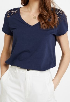 Femme portant un T-shirt bleu marine col V avec des détails en dentelle aux épaules et un pantalon blanc taille haute, mains dans les poches.