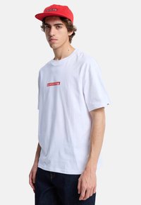 Hvid bomulds t-shirt med et rødt rektangulært logo på brystet. Parret med en rød cap med et lille logo. Enkel, afslappet design.