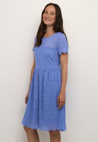 Blå blondekjole med korte ærmer, bølget kant og tætsiddende bodice. Stofmet er mønstret med blomster og falder ned i en udvidet nederdel.