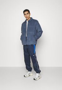 Blå ruskindsjakke med knappelukning, båret over en grå skjorte. Navy trackpants med blå sidelister og sølvfarvede sportssko.