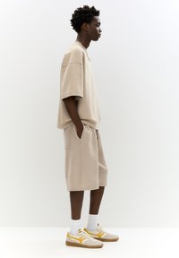 Tricou oversized bej și șorturi asortate, ambele din bumbac. Pantofii sunt bej cu accente galbene. Modelul stă în profil.