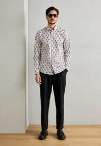 Λευκό πουκάμισο με κουμπιά, διακοσμημένο με πολύχρωμο λουλουδάτο σχέδιο, συνδυασμένο με μαύρο παντελόνι και χοντρές μαύρες παπούτσια. Το μοντέλο φορά ηλιόυαθιο.