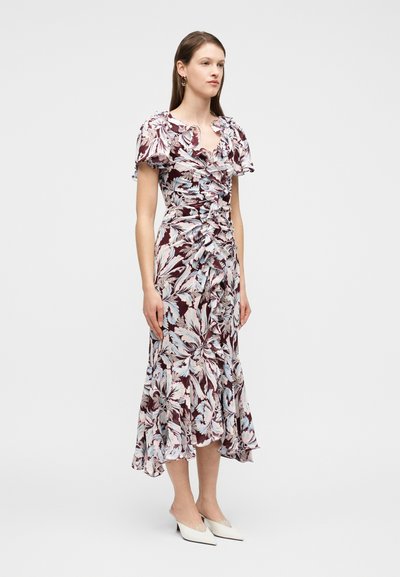 Γυναίκα όρθια, φορώντας μωβ midi φόρεμα με λουλουδάτο σχέδιο και βολάν, με κοντά μανίκια και λευκά γυναικεία παπούτσια με ψηλό τακούνι και μυτερή μύτη, σε απλό φόντο.