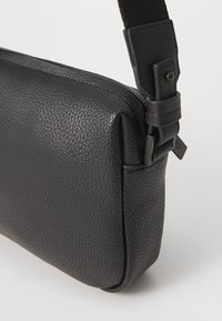 Borsa a spalla in pelle nera con superficie testurizzata, forma rettangolare e tracolla regolabile. Presenta una chiusura a zip e dettagli in metallo.