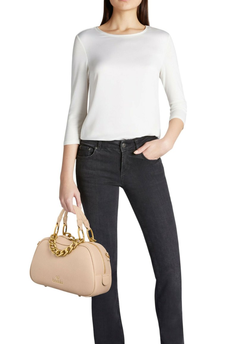 Borsa beige con superficie texturizzata, manico a catena dorata e chiusura con zip, abbinata a una maglietta bianca e jeans neri.