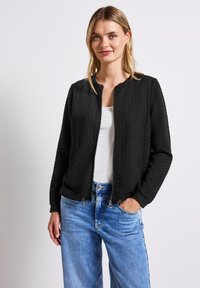 Černá prošívaná bomber bunda s kulatým výstřihem, zapínáním na zip a dlouhými rukávy. V kombinaci s světle modrými širokými džíny.
