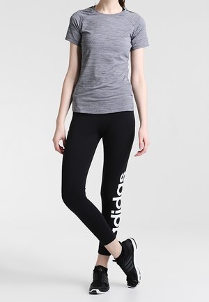 Nainen pukeutuneena tiukkaan harmaaseen lyhythihaiseen paitaan, mustiin Adidas-legginseihin, joissa valkoinen logo, ja mustiin urheilukenkiin, seisoo jalkoja ristissä.