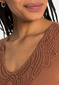 Blusa marrom com decote em V e detalhes em renda intrincados ao redor do decote; tecido suave e texturizado.