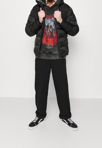 Giacca a vento nera con finitura testurizzata, t-shirt grigia con un design rosso, pantaloni neri e sneakers nere con dettagli bianchi.