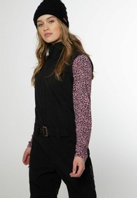 Čierna bezrukávová zipsová vesta cez ružový top s dlhými rukávmi a čiernymi leopardími škvrnami. Vesta má opasok a subtílne detaily z kovu.