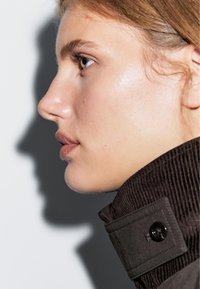 Brązowy kołnierz z kordurojowym wykończeniem i akcentem w postaci guzika, zbliżenie na twarz ukazujące gładką cerę, podkreślające kształt nosa i ust.