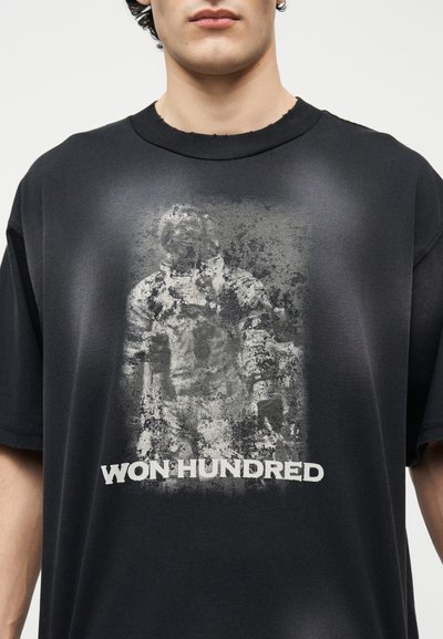 Άνδρας που φοράει μαύρο μπλουζάκι με ξεθωριασμένο σχέδιο ενός αστροναύτη και το κείμενο "WON HUNDRED" κάτω από την εικόνα.