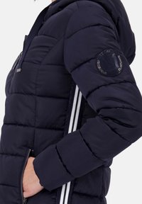 Marineblau gepolsterte Jacke mit Kapuze, mit einem gesteppten Design, zwei seitlichen Taschen und weißen Streifen an den Seiten. Logopatch am Ärmel.