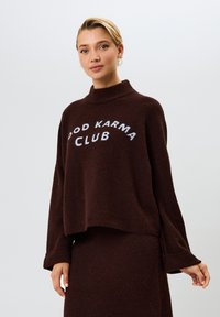 Ruskea, lyhyt neulepaita pehmeästä materiaalista. Ominaisuuksiin kuuluu korkea kaulus ja leveät hihat. Rinnassa on valkoisella teksti "GOOD KARMA CLUB".