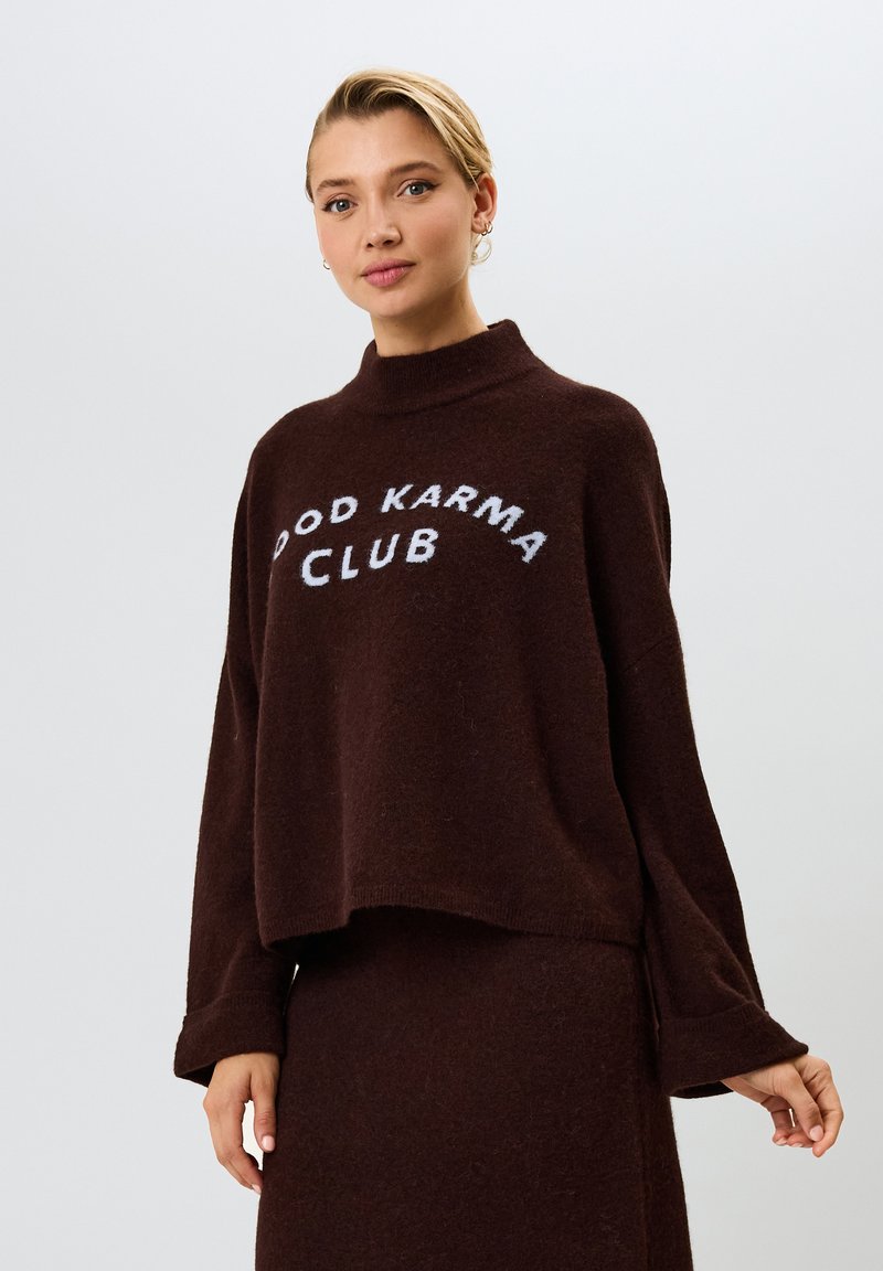 Ruskea, lyhyt neulepaita pehmeästä materiaalista. Ominaisuuksiin kuuluu korkea kaulus ja leveät hihat. Rinnassa on valkoisella teksti "GOOD KARMA CLUB".