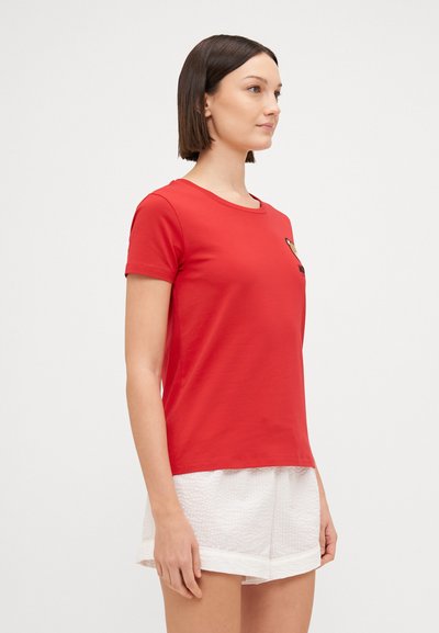 Κόκκινο κοντομάνικο μπλουζάκι με στρογγυλή λαιμόκοψη και διακοσμητική ετικέτα στο στήθος, συνδυασμένο με άσπρες υφ ασμάτινες σορτς.