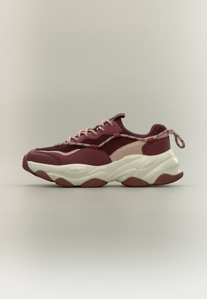 Baskets épaisses avec dessus en mesh et cuir bordeaux, rose et beige, semelle blanche épaisse, lacets roses et languette au talon sur un fond neutre.