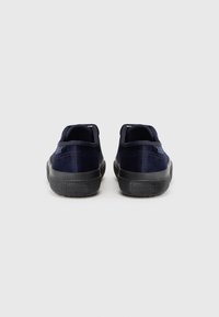 Pantofi slip-on din catifea bleumarin cu o talpă din cauciuc negru texturat și o bandă de prindere subtilă la spate pentru a facilita îmbrăcarea.