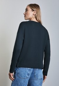 Schwarzes Langarmshirt aus glattem Stoff, mit lockerem Schnitt und seitlichen Schlitzen, kombiniert mit hellblauen Jeans.