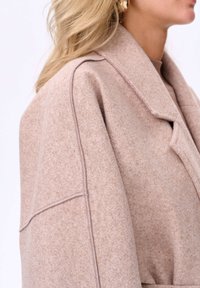 Jasnoróżowy, oversizeowy wełniany płaszcz z rozciętym kołnierzem i zaokrąglonymi ramionami. Charakteryzuje się widocznym przeszyciem i miękką, teksturowaną powierzchnią.