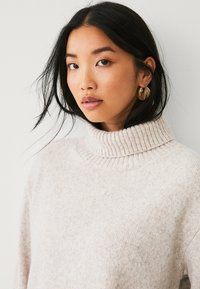 Maglione a collo alto beige chiaro con colletto a coste, texture morbida e vestibilità comoda. Modella orecchini circolari di grandi dimensioni.