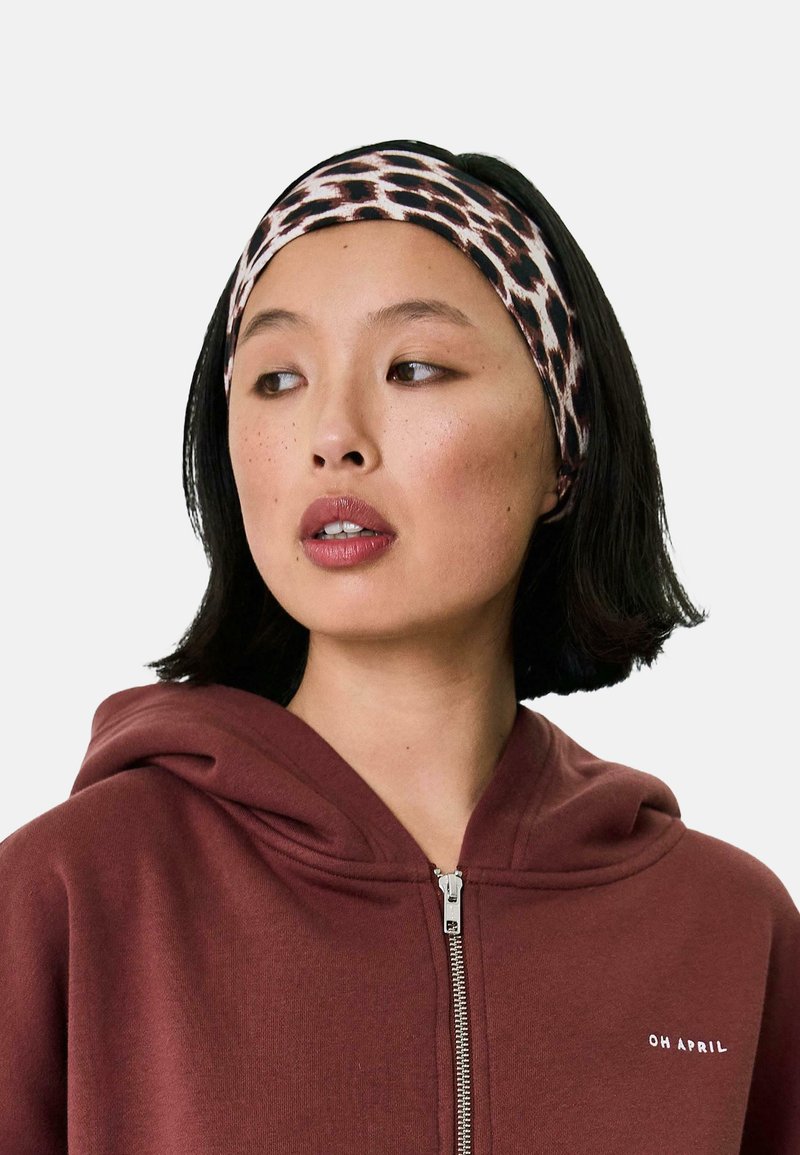 Leopardų rašto galvos juosta, pagaminta iš tamprios medžiagos, su šviesių ir tamsių spalvų raštu, dėvima su bordo spalvos zip-up džemperiu.