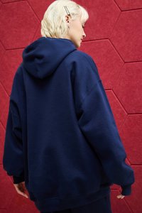Felpa oversize blu navy realizzata in materiale morbido, con un grande cappuccio e polsini a coste. Sfondo in tessuto rosso esagonale.