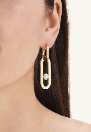 Gold rectangular drop earring with small crystals and a central pearl, worn on a woman's ear with dark hair.