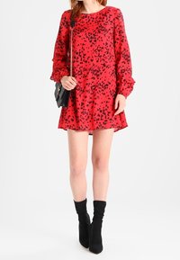 Rotes Kleid mit schwarzen Punkten, langen Ärmeln und ausgestelltem Saum. Getragen mit schwarzen kniehohen Stiefeln und einer kleinen Umhängetasche.