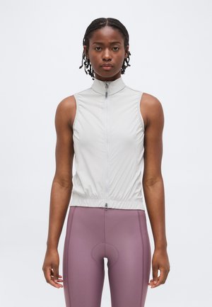 Fonott hajú nő világosszürke ujjatlan zipzáras mellényben és lila magas derekú kerékpáros leggingsben, egyszínű fehér háttér előtt áll.