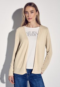 Beige kashmirtröja med öppen front, långa ärmar, texturerad tyg. Bärs över en vit skjorta med texten "NATURE & REVIVAL" tryckt i svart. Blå jeans.