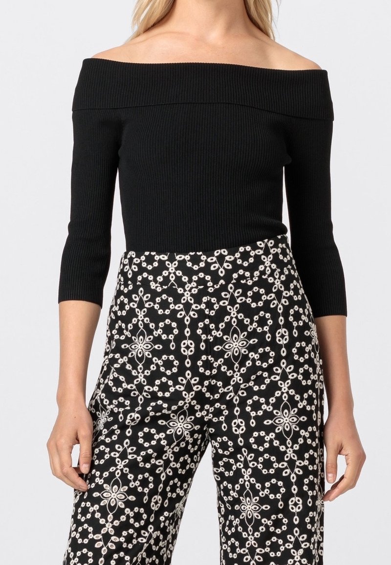 Femme portant un haut côtelé noir décolleté épaule avec des manches trois-quarts et un pantalon noir taille haute avec un motif géométrique floral blanc.