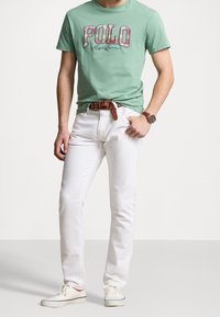 Green cotton t-shirt with "POLO" graphic, paired with white denim jeans. Brown woven belt and white sneakers complete the look.