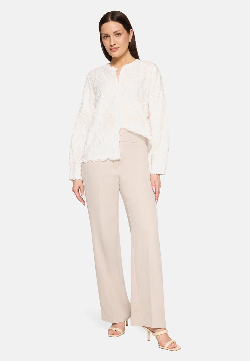 Femme portant un chemisier blanc à manches longues texturé et un pantalon large beige avec des talons ouverts beige, debout devant un fond uni.