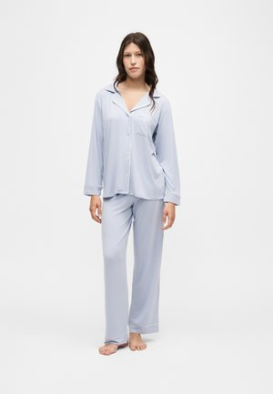 Femme debout pieds nus portant un haut de pyjama bleu clair à manches longues avec des boutons et un pantalon de pyjama ample assorti sur fond blanc uni.