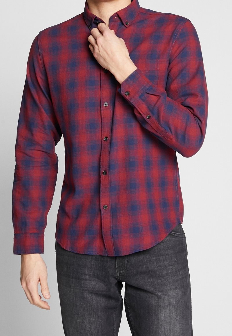 Homme portant une chemise à carreaux rouge et bleu foncé à manches longues boutonnée, avec un jean gris foncé, ajustant le col d'une main.