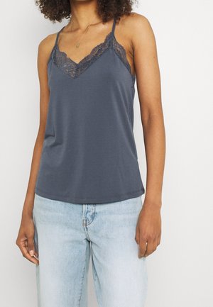 Femme portant un caraco gris foncé bordé de dentelle et un jean taille haute bleu clair, debout devant un fond uni.