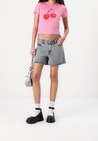 Kvinna iklädd en rosa kortärmad t-shirt med röda körsbär, grå denimshorts, vita strumpor, svarta skor och hållande en silverhandväska.