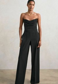 Svart strapless jumpsuit med ett samlat halsringning, vida ben och sidofickor. Modellen bär ett guldhalsband mot en neutral bakgrund.
