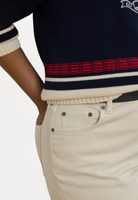 Marineblå strikkegenser med røde og kremfargede striper, kombinert med kremfargede jeans med metallknappdetaljer og en sømløs livlinje.