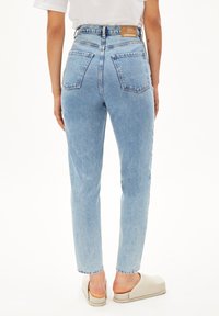 Jean taille haute bleu clair avec une coupe droite, comportant deux poches arrière et une étiquette beige au niveau de la taille. Associés à des sabots beiges.