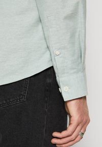 Camicia verde chiaro con colletto, texture sottile e polsini con bottoni, abbinata a pantaloni in denim scuro.