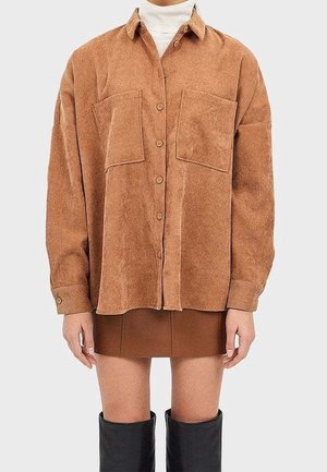 Person iført en løs brun skjorte med store brystlommer, en kort brun nederdel og sorte knælange støvler.